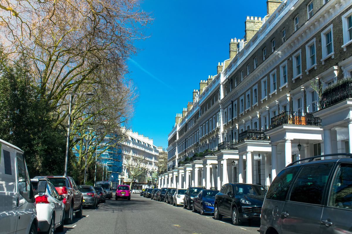 A busy residential road in London with Townhouses lining one side of the road and a park on the other